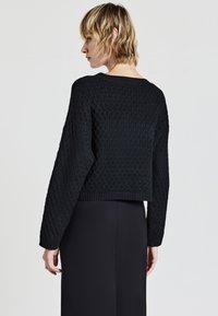 Donna con capelli biondi corti che indossa un maglione nero a maglia con texture e una gonna nera, mostrata di spalle su uno sfondo semplice.