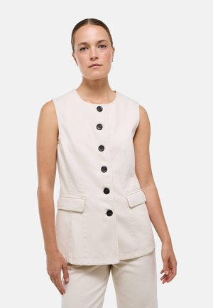 Femme aux cheveux tirés en arrière portant un gilet beige sans manches boutonné et un pantalon assorti sur un fond clair uni.