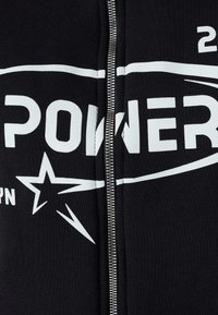 Felpa nera con zip realizzata in cotone, con testo stampato bianco e grafica di una stella. Cerniera argentata che scorre lungo il centro.