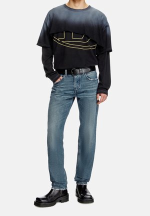 Homme portant un t-shirt à manches longues noir superposé avec un graphisme jaune, un jean bleu, une ceinture noire et des bottes en cuir noires, debout avec une main dans la poche.