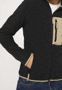 Mężczyzna w czarnej polarowej kurtce zapinanej na zamek z beżowymi wykończeniami i kieszenią na piersi, z ręką w kieszeni bocznej, w połączeniu z niebieskimi dżinsami.