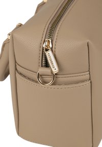 Bézs bőrtáska sarok arany cipzárral és húzófüllel, melybe be van gravírozva a "VIVISENCE" felirat, oldalt pedig egy arany karika található.