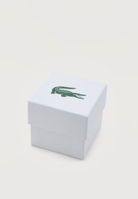 Λευκό υφασμάτινο τετράγωνο κουτί με πράσινο λογότυπο κροκοδείλου στην κορυφή, τοποθετημένο σε λείο λευκό υπόβαθρο.