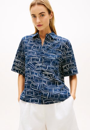 Kvinde iført en mørkeblå kortærmet skjorte med hvide abstrakte linjer og hvide bukser, stående med hænderne i lommerne.