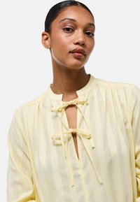 Blouse jaune clair avec des rayures verticales, détails froncés près du col et une découpe en trou de serrure maintenue par des liens. Texture lisse.