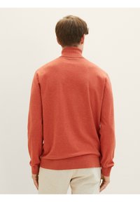 Rode gebreide trui met een hoge kraag, geribbelde manchetten en onderrand. Zachte textuur en een iets losse pasvorm. Draag het over lichtgrijze onderstukken.