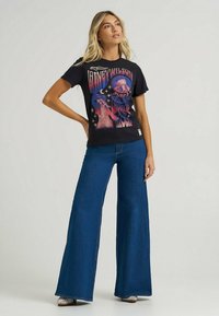 Camiseta gráfica negra con estampado colorido y texto, combinada con pantalones vaqueros de pierna ancha en azul oscuro y zapatos plateados con purpurina.