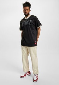Cămașă scurtă din plasă neagră cu logo-ul "FUBU" în alb, cu detalii crem, asortată cu pantaloni crem și adidași roșii și albi.
