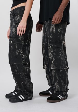 Πλάγια όψη δύο ατόμων που φορούν μαύρο παντελόνι καρό cargo με όξινη πλύση και μαύρα αθλητικά παπούτσια με λευκές ρίγες, στέκονται μπροστά σε απλό φόντο.