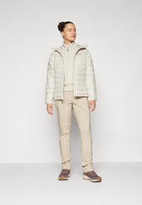 Lett, fyldig kremjakke med glidelås, parret med en beige jakke med glidelås og lyse beige bukser. Brune tursko med teksturerte såler.