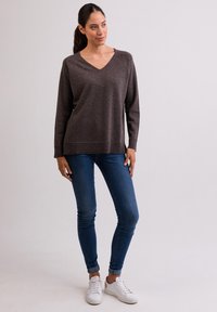 Brun v-ringad tröja i mjukt material, kombinerad med blå smala jeans och vita sneakers, visad på en stående modell mot en enkel bakgrund.