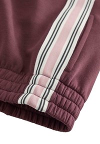 Bordeauxfarbene Sweatpants mit elastischem Bund, versehen mit rosa und schwarzen gestreiften Akzenten an der Seite. Glatter Stoff mit lockerem Schnitt.