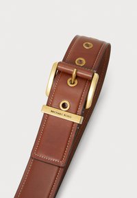Barna bőrövezett öv arany fém csattal és arany díszítéssel, amelyen a "MICHAEL KORS" felirat olvasható. Feltűnő varrásokkal és fémbetétekkel rendelkezik.