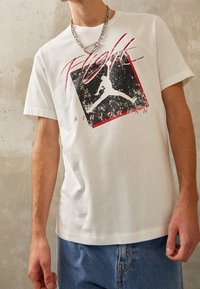 Persona vestida con una camiseta blanca Air Jordan Flight con logo negro y rojo y unos jeans de mezclilla azul claro, contra una pared beige texturizada.