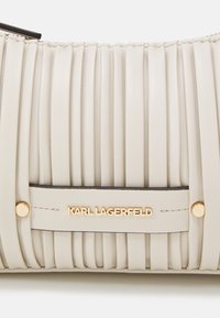 Sac en cuir beige avec des plis verticaux, présentant un logo et des accents dorés. Texture lisse avec une forme épurée et structurée.
