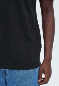 Pull-over noir en maille lisse, avec un ourlet côtelé et une étiquette sur le côté, porté avec un jean en denim bleu clair.