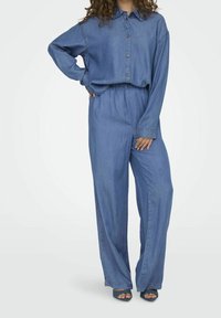 Woman wearing loose-fitting, button-up denim shirt tucked into high-waisted, wide-leg denim pants with blue heeled sandals.