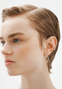 Un pendiente en forma de lágrima de plata cuelga de la oreja del modelo. El cabello corto está peinado hacia atrás. La piel está clara con sutiles pecas. Diseño minimalista.