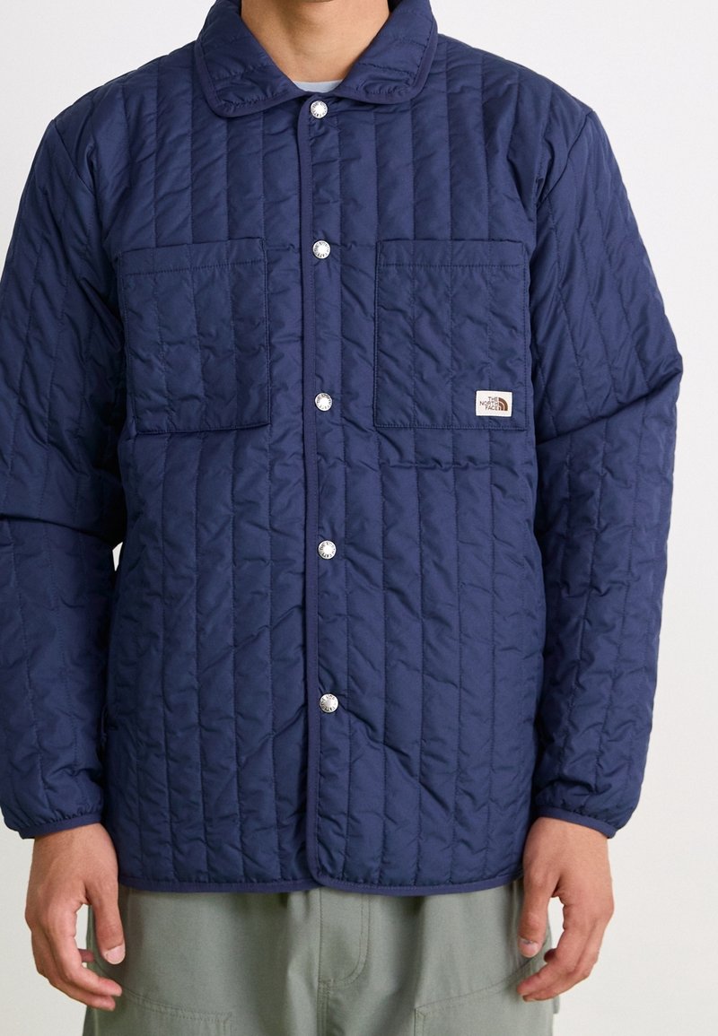Personne portant une veste matelassée bleu marine avec des boutons-pression et deux poches poitrine, dont une poche avec un petit patch logo de la marque.