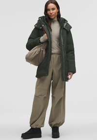 Zelená prošívaná bunda s kapucí, béžový top, volné světlé béžové kalhoty a černé boty. Taška je krémová s nařaseným otvorem.