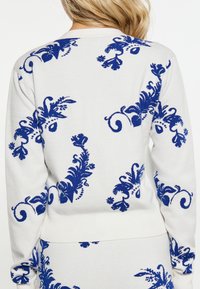 Biały sweter z niebieskimi wzorami kwiatów i winorośli noszony przez osobę zwróconą plecami, pokazującą górną część pleców i część spódnicy.