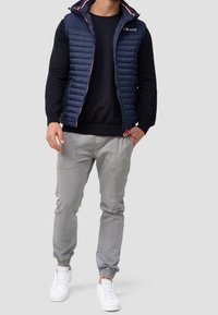 Smanicato imbottito blu navy con cappuccio e dettagli rossi a contrasto, indossato sopra un maglione nero, abbinato a pantaloni jogger grigi e sneakers bianche.