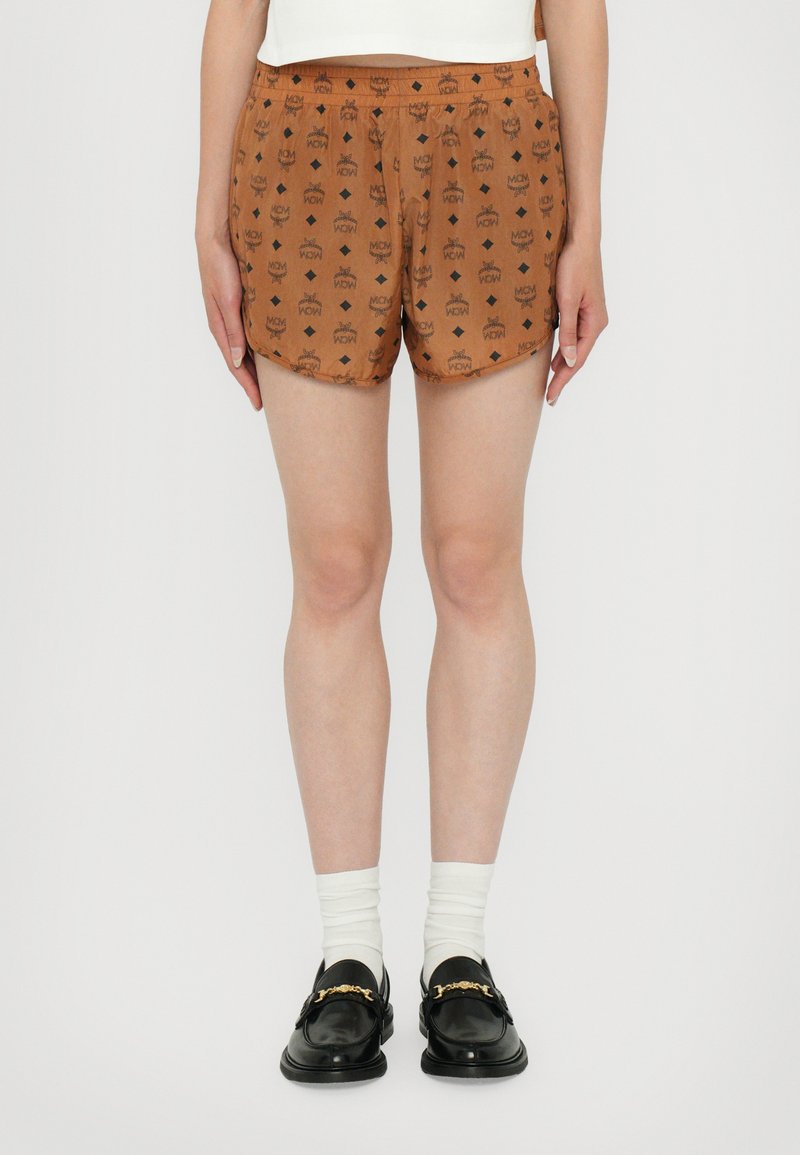 Braune Shorts mit einem sich wiederholenden Kronenmuster, einem elastischen Bund und einem leichten Stoff, kombiniert mit schwarzen Slippern und weißen Socken.