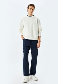 Sudadera de algodón blanca con cuello de canalé, combinada con pantalones de chándal texturizados en azul marino. El modelo lleva zapatillas blancas. Ajuste sencillo y relajado.