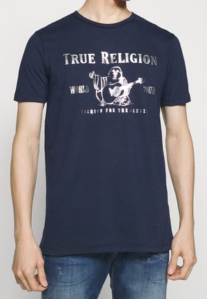 T-shirt blu navy con testo argento "TRUE RELIGION WORLD TOUR" e grafica di un Buddha che suona la chitarra, indossata con jeans blu.