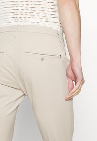 Pantalon beige clair, coupe slim, doté de deux poches arrière, fermetures à boutons et d'un tissu lisse et texturé.