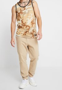Débardeur en maille beige clair avec un motif camouflage, associé à un pantalon jogging beige clair et des baskets blanches. Comprend une étiquette logo sur le débardeur.