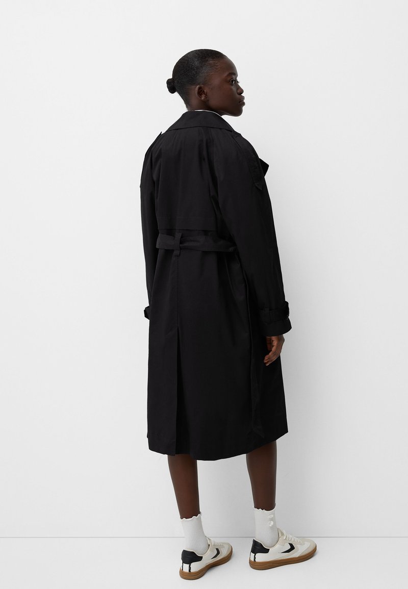 Kobieta o ciemnej karnacji ubrana w czarny trencz, białe skarpetki sięgające kostek i białe tenisówki, stoi przodem do białej ściany.