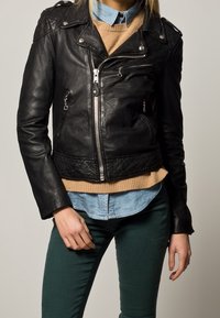 Veste de moto en cuir noir avec fermeture éclair à l'avant, ourlet inférieur matelassé, quincaillerie argentée et deux poches latérales portée par-dessus un pull beige et une chemise en denim.