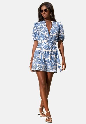 Woman wearing sunglasses and a white and blue patterned belted romper with puff sleeves and white strappy sandals, walking forward.