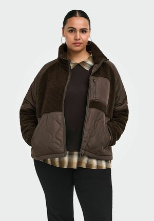 Femme aux cheveux foncés portant une veste matelassée et en polaire marron par-dessus une chemise à carreaux et un pantalon noir, debout les mains dans les poches.