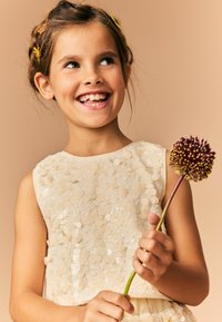 De jurk heeft een mouwloos ontwerp met pailletten in crèmekleuren. Het kind houdt een bloem vast met een ronde paarse bloemkop en een groene steel tegen een beige achtergrond.