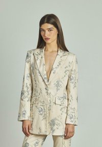 Completo in lino ricamato floreale beige con motivi blu. Caratterizzato da un colletto a punta, bottoni frontali e due tasche anteriori. Vestibilità su misura.