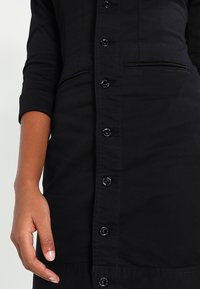 Czarna sukienka zapinana na guziki o prostym kroju, z długimi rękawami, przednią listwą z czarnymi guzikami oraz małą kieszonką na piersi.