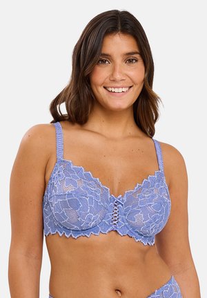 Femme souriante portant un soutien-gorge en dentelle violet clair avec motif floral et bretelles ajustables sur un fond uni.
