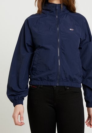 Kobieta ubrana w granatową kurtkę na zamek z elastycznymi mankietami i dolnym brzegiem, w zestawieniu z czarnymi spodniami na jednolitym jasnym tle.