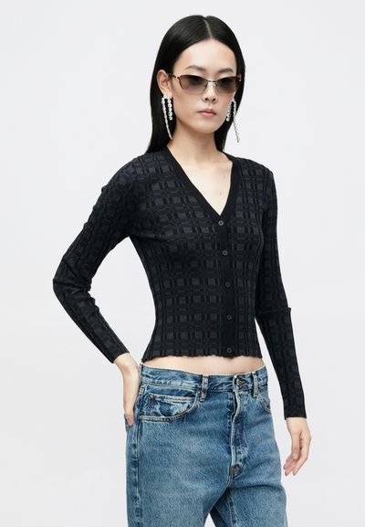 Cardigan negru, scurt, cu mâneci lungi, cu o textură cu pătrățele și guler adânc în formă de V; are închidere cu nasturi și margine ondulată. Purtați peste blugi albaștri.