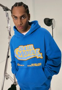 Blå huvtröja med gul grafisk text "Still Moving" och mindre text nedanför. Har en känguruficka och dragsko i huvan.