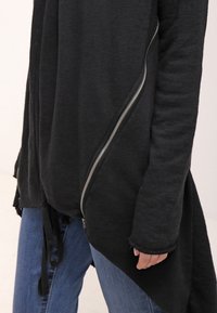 Černý zipový top s drapovaným designem, s dlouhým asymetrickým lemem a strukturovanou tkaninou. V kombinaci s modrými džíny.