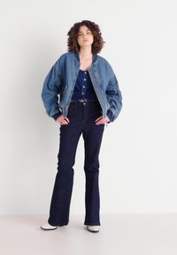 Denim bombarder jakna, svijetloplava, s rebrastim manžetama, preko tamnoplave košulje s dugim rukavima i tamnih traperica s flared krojem, u kombinaciji s bijelim čizmama.