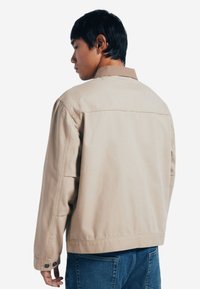 Giacca di jeans beige con colletto marrone, maniche lunghe, polsini abbottonati e taglio dritto. Mostrata da dietro, indossata sopra dei jeans blu.