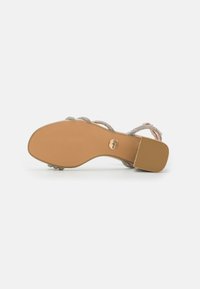 Sandal con una suela de color marrón claro y múltiples tiras trenzadas entrelazadas que presentan acentos metálicos, mostrando un tacón plano y una textura suave.