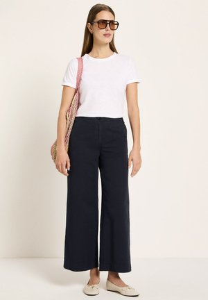 Woman wearing oversized brown sunglasses, white short-sleeve shirt, wide-leg black pants, beige flats, and carrying a pink knit shoulder bag.