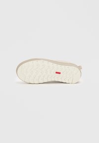 Suela de zapatilla beige con un patrón de trato profundo y un pequeño logo rectangular rojo en el centro, exhibido sobre un fondo blanco liso.