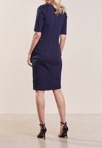 Žena s blond vlasy, oblečená v přiléhavých námořnických šatech ke kolenům a černých lodičkách na vysokém podpatku s náramkem kolem kotníku, stojící na neutrálním pozadí.