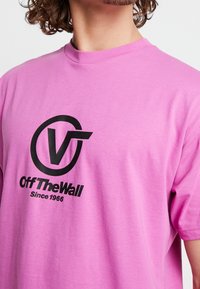 Vaaleanpunainen puuvillainen t-paita, jossa on suuri musta logo, jossa on pyöreä muotoilu ja teksti "Off The Wall Since 1966." Lyhyet hihat.